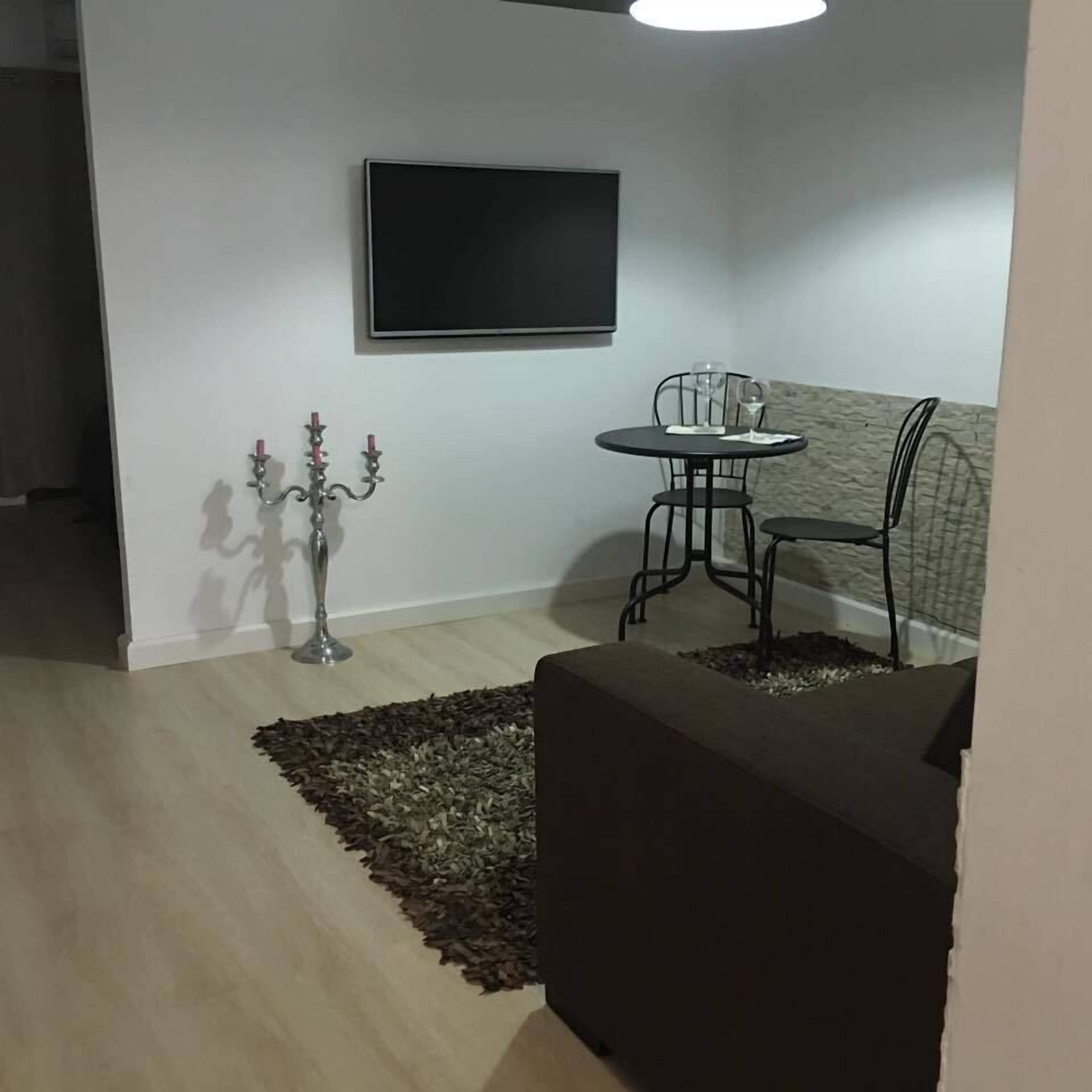 Una sala de estar con una televisión de pantalla plana montada en la pared, una mesa redonda con dos sillas, un candelabro decorativo y una alfombra estampada.