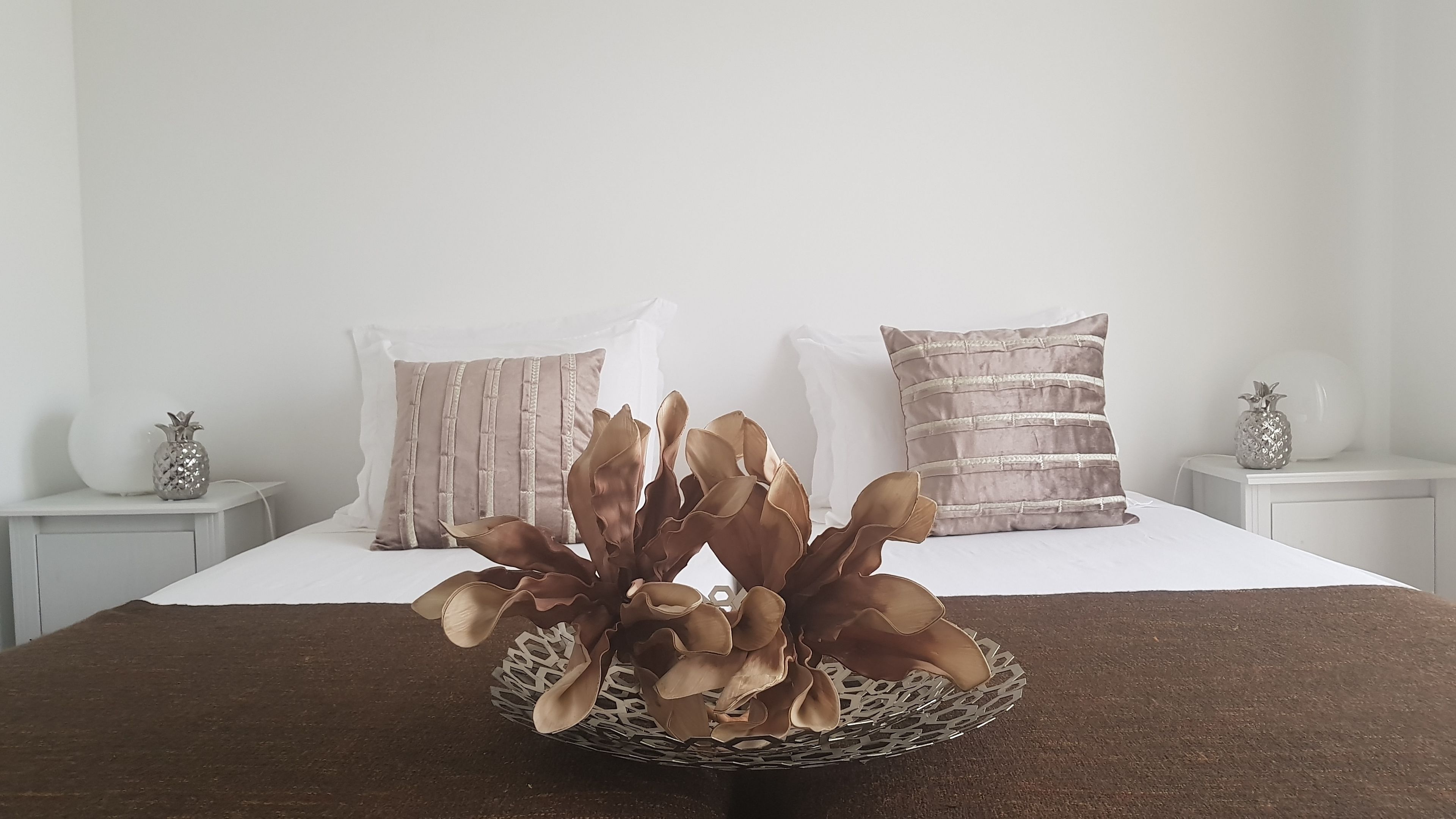 Een bed met een witte spreideken, twee decoratieve kussens, een tafeltje met een vaas in de vorm van een ananas, en een decoratieve schaal met gedroogde bloemen.