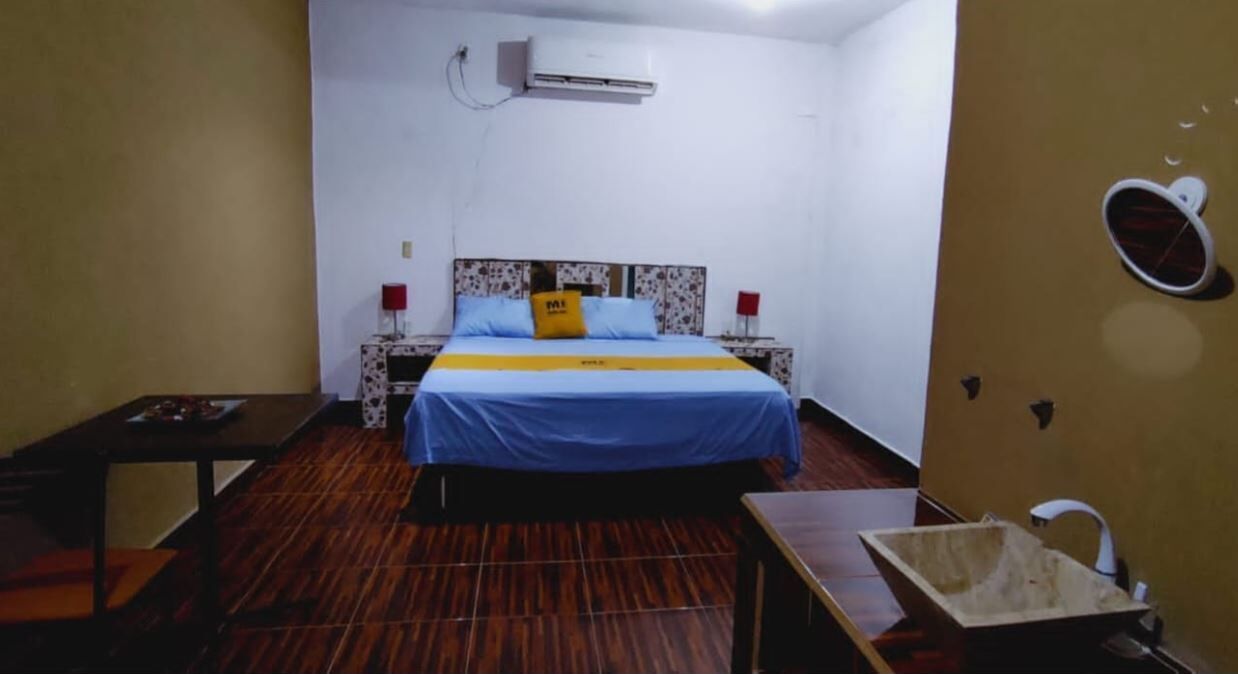 Una habitación con cama, una mesita, una silla, un equipo de aire acondicionado y un espejo instalado en la pared.