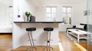 A modern kitchen with a central island, two bar stools, and a dining area with a white table and sofa. - Modern Apartment In The City Center (Bergen)