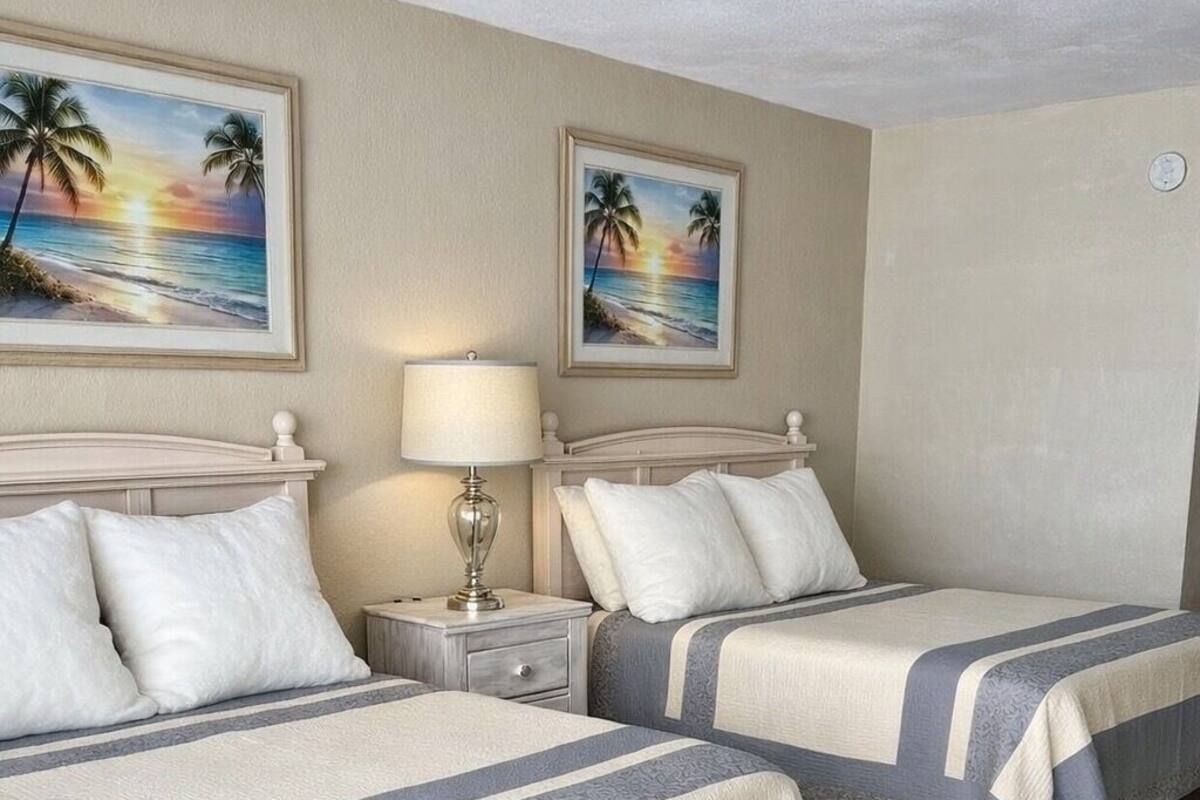 A hotel room with two beds, a nightstand with a lamp, and two framed sunset beach paintings on the wall.