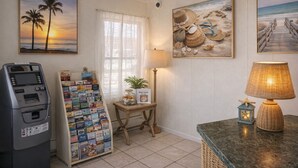 A room with a beach-themed wall art, a small table with a lamp, a magazine rack, and a cash machine.