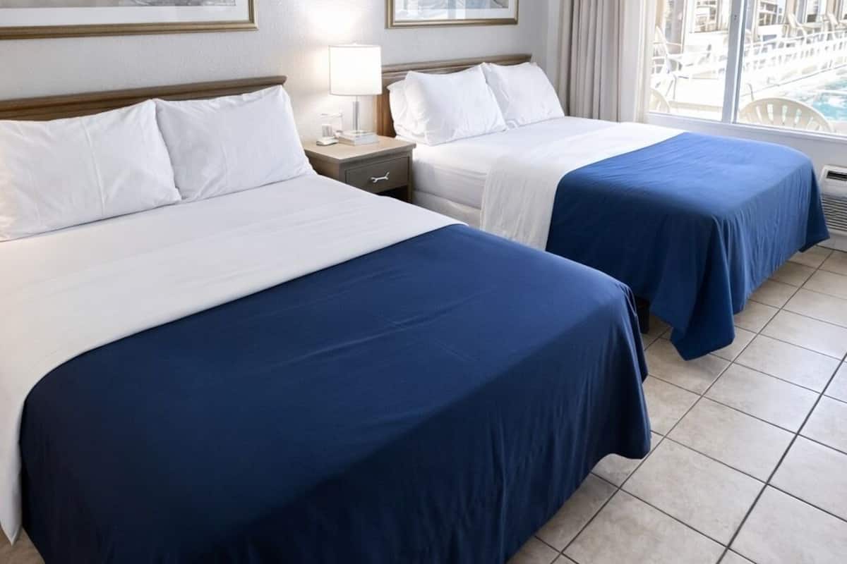 A hotel room with two beds, a nightstand, a lamp, and a large window with a view of a pool and outdoor area.