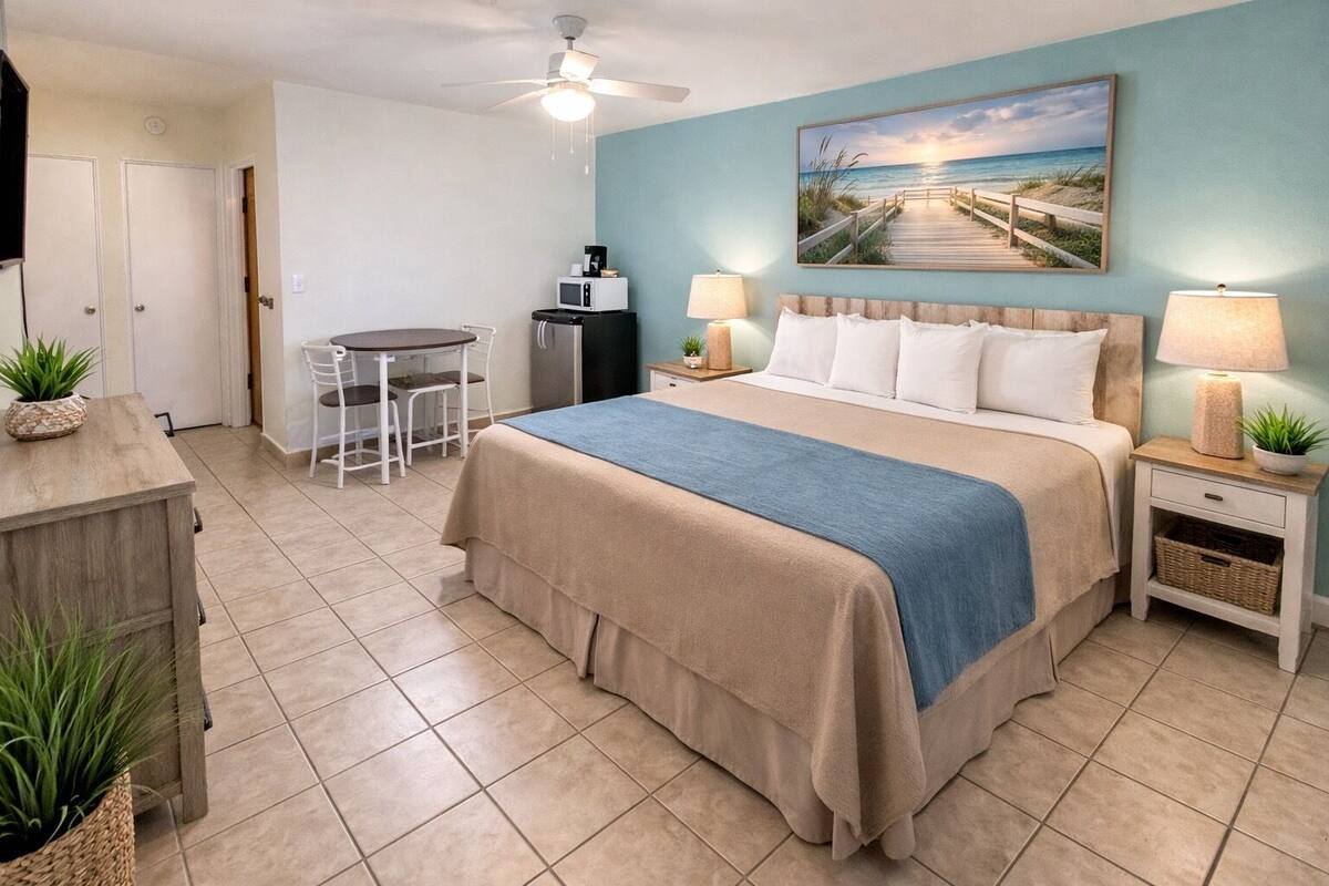 A bedroom with a bed, bedside lamps, a desk, a chair, a refrigerator, a microwave, and a painting of a beach scene.