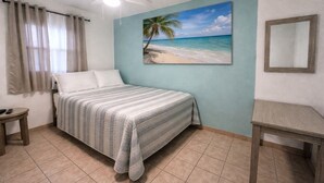 A bedroom with a bed, a ceiling fan, a painting of a beach, a mirror, and a small table.