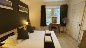 Une chambre d’hôtel équipée d’un lit, d’un bureau, d’une chaise, d’un ventilateur sur pied et d’une fenêtre avec des rideaux.