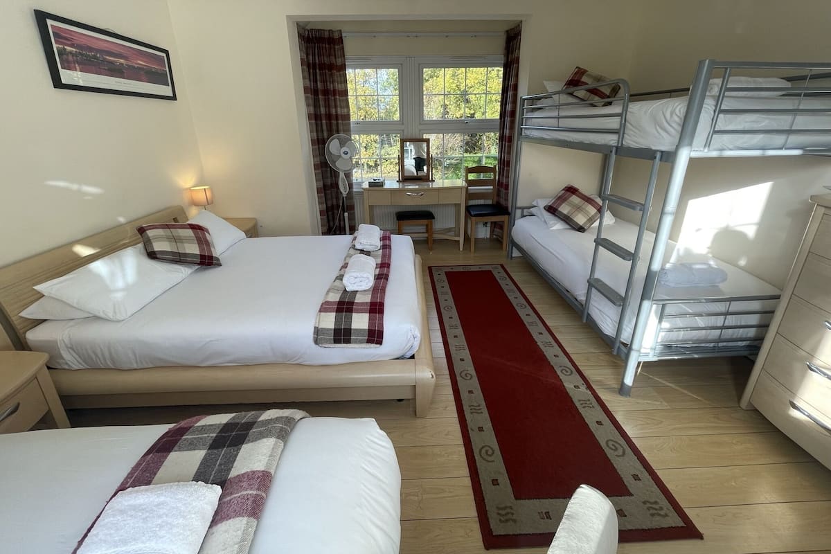 Quarto de hotel com uma cama grande, uma cama menor, uma escrivaninha com cadeira, uma beliche e uma janela com cortinas.