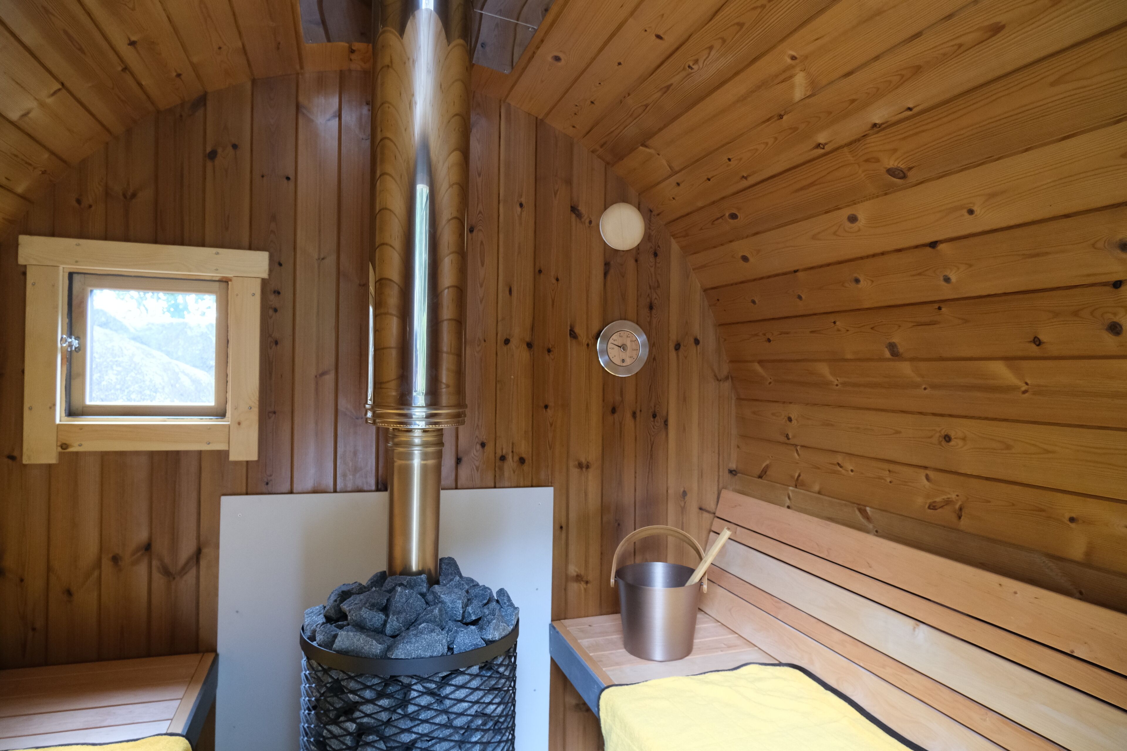A wooden cabin interior with a wood stove, a window, and a metal bucket on a wooden table.