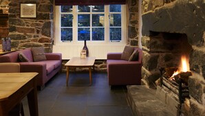 Una habitación acogedora con paredes de piedra, chimenea y un amplio ventanal con vistas a árboles. Hay dos sofás y una mesa de madera.