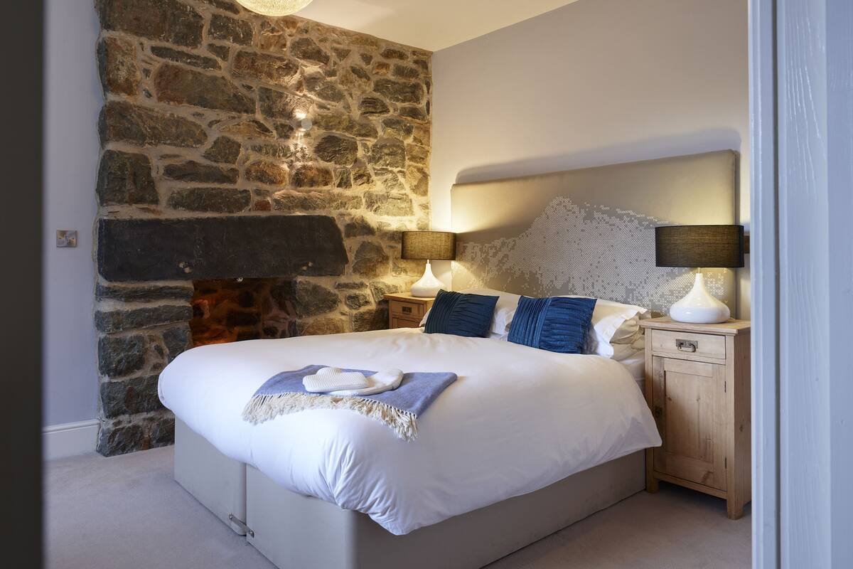 Een slaapkamer met een stenen muur, een bed met witte lakens, een hanglamp en een nachtkastje met een lamp.