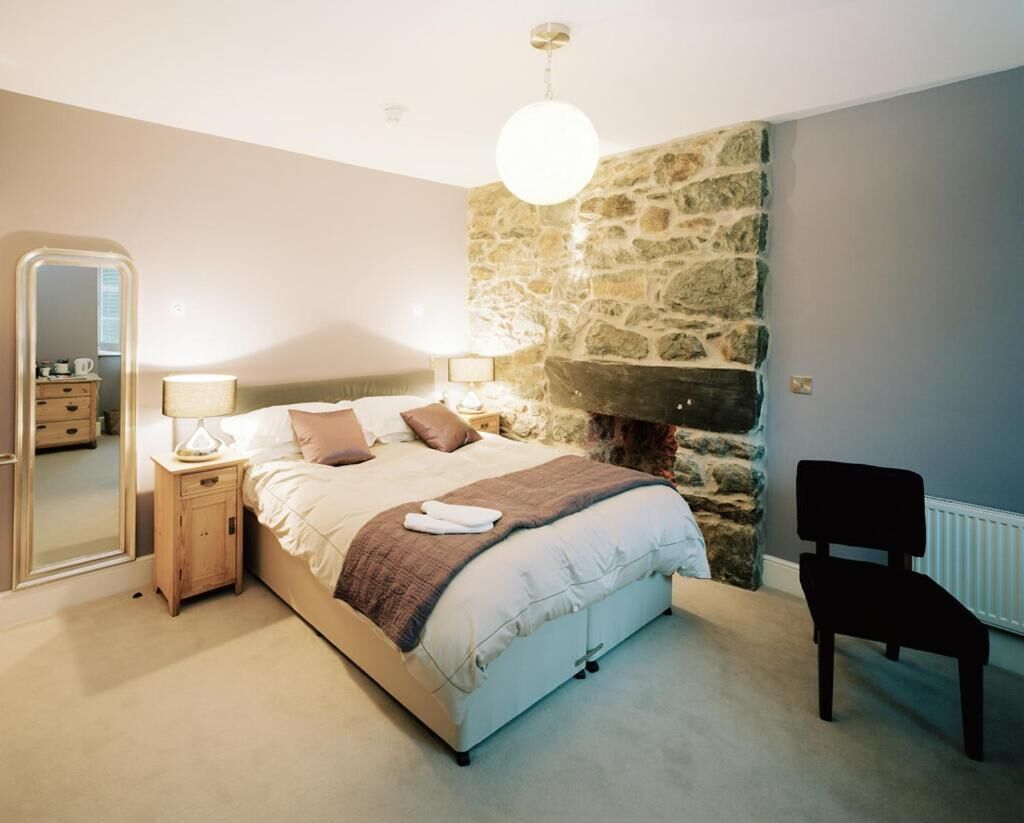A bedroom with a stone wall, a bed with pillows, a nightstand with a lamp, and a chair.