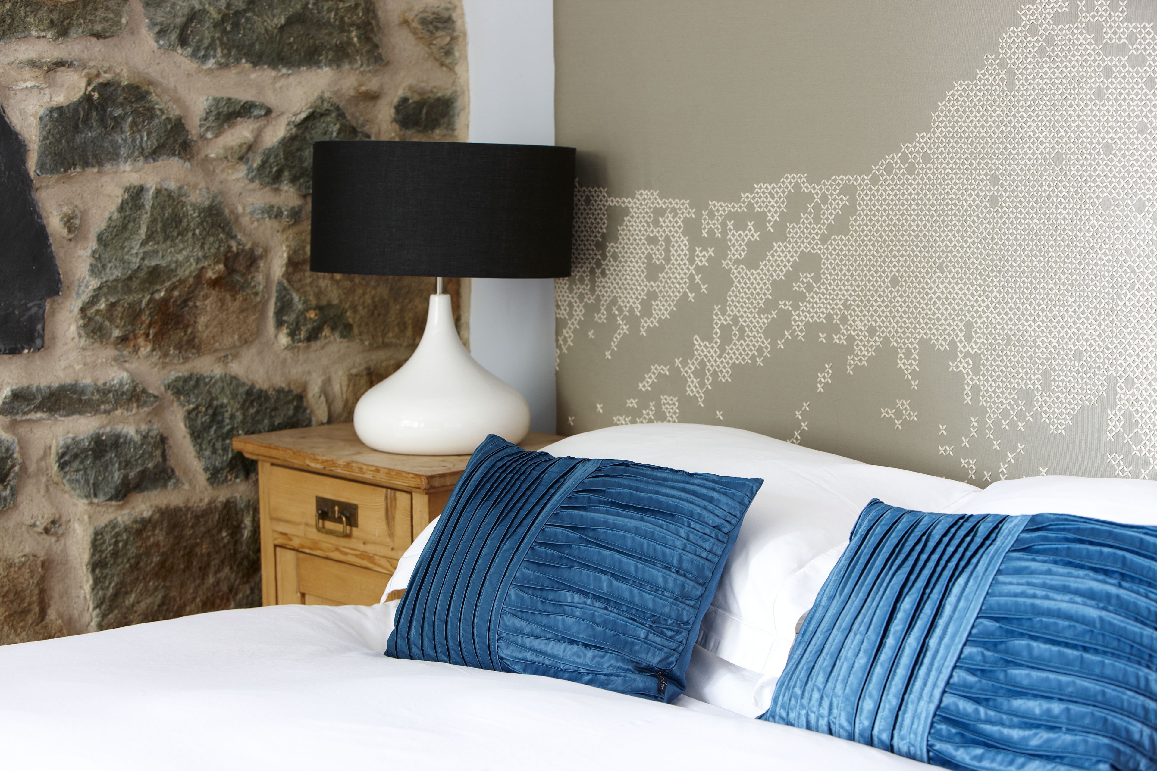 A bed with blue pillows, a white lamp on a wooden nightstand, and a stone wall.