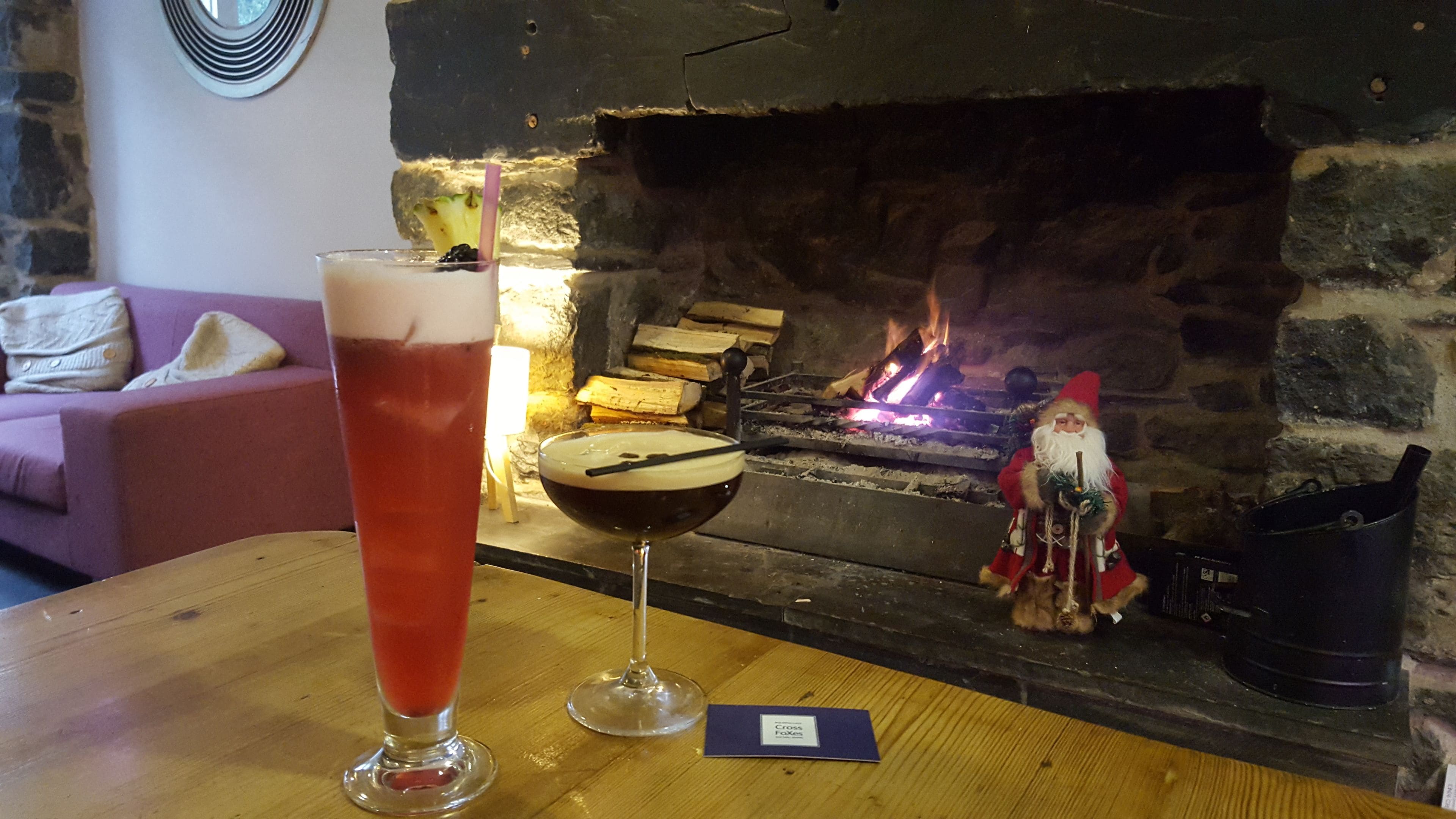 A table with a glass of beer and a cocktail, a small card, and a fireplace with logs and a Santa figurine.