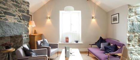 Un salón acogedor con pared de piedra, una mesa de madera, un sofá morado y una ventana con luz natural.