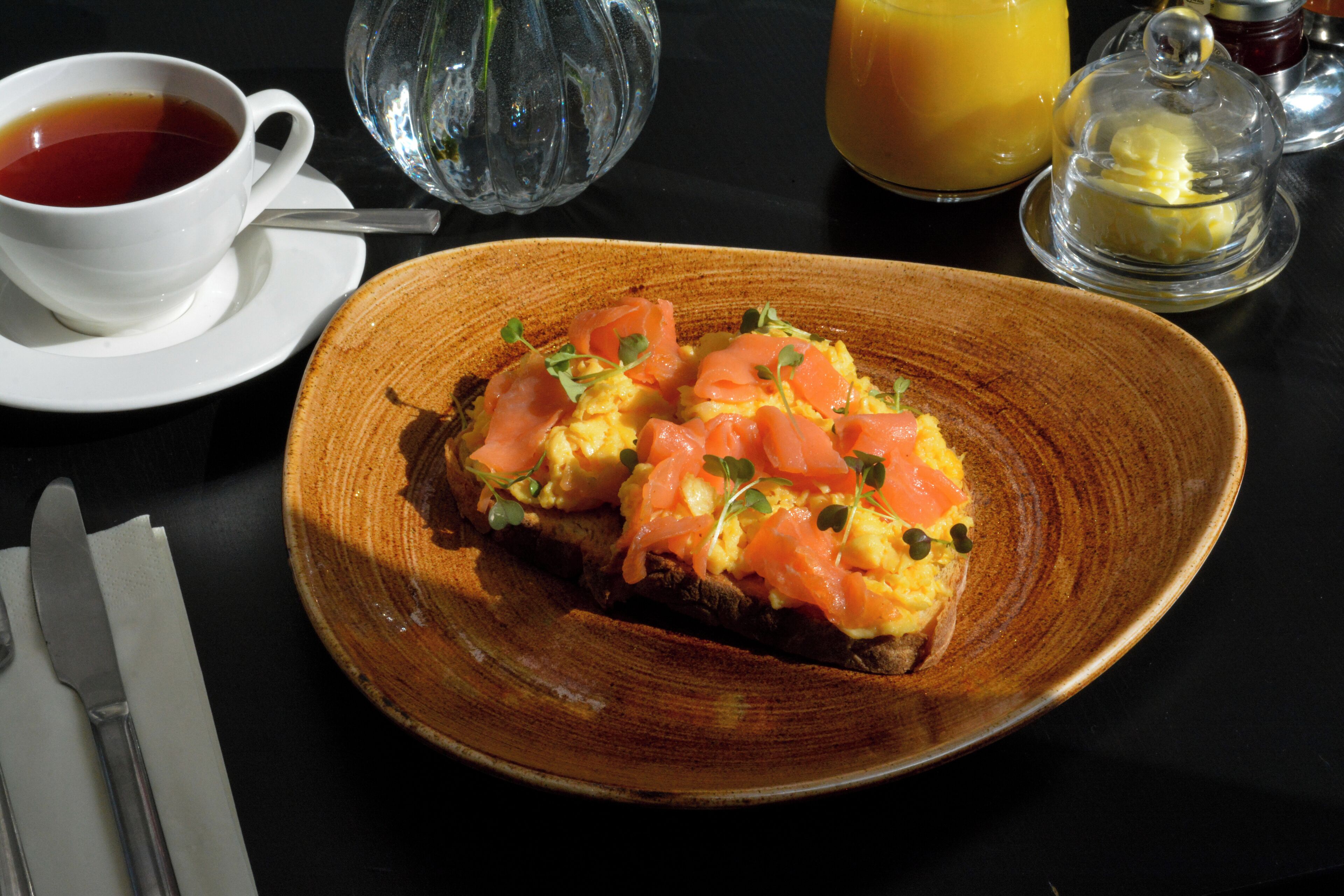 A breakfast plate with eggs and smoked salmon on toast, a cup of tea, and a glass of orange juice on a table.