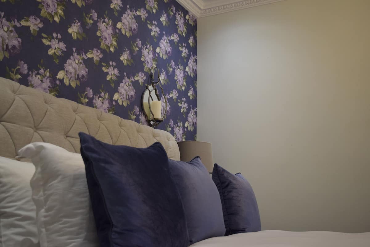 A bedroom with a tufted headboard, a bed with white and dark blue pillows, a wall with floral wallpaper, and a bedside lamp.