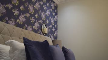 Camera da letto con testiera trapuntata, un letto con cuscini bianchi e blu scuro, una parete con carta da parati a motivi floreali e una lampada da comodino.