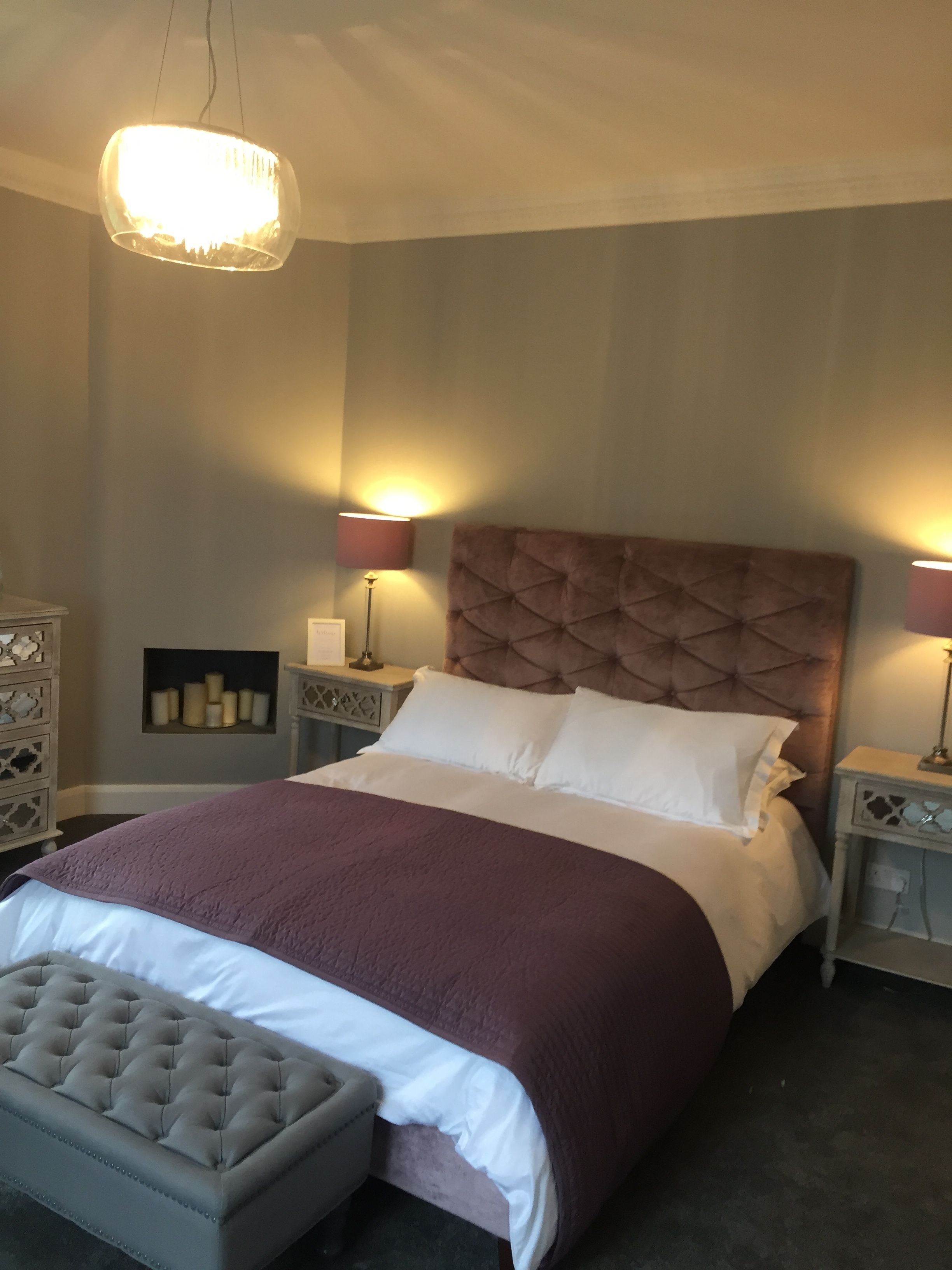 Een net opgemaakt bed met een gestoffeerd hoofdeinde, twee nachtlampjes, een decoratief wandobject en een gestoffeerde bank.