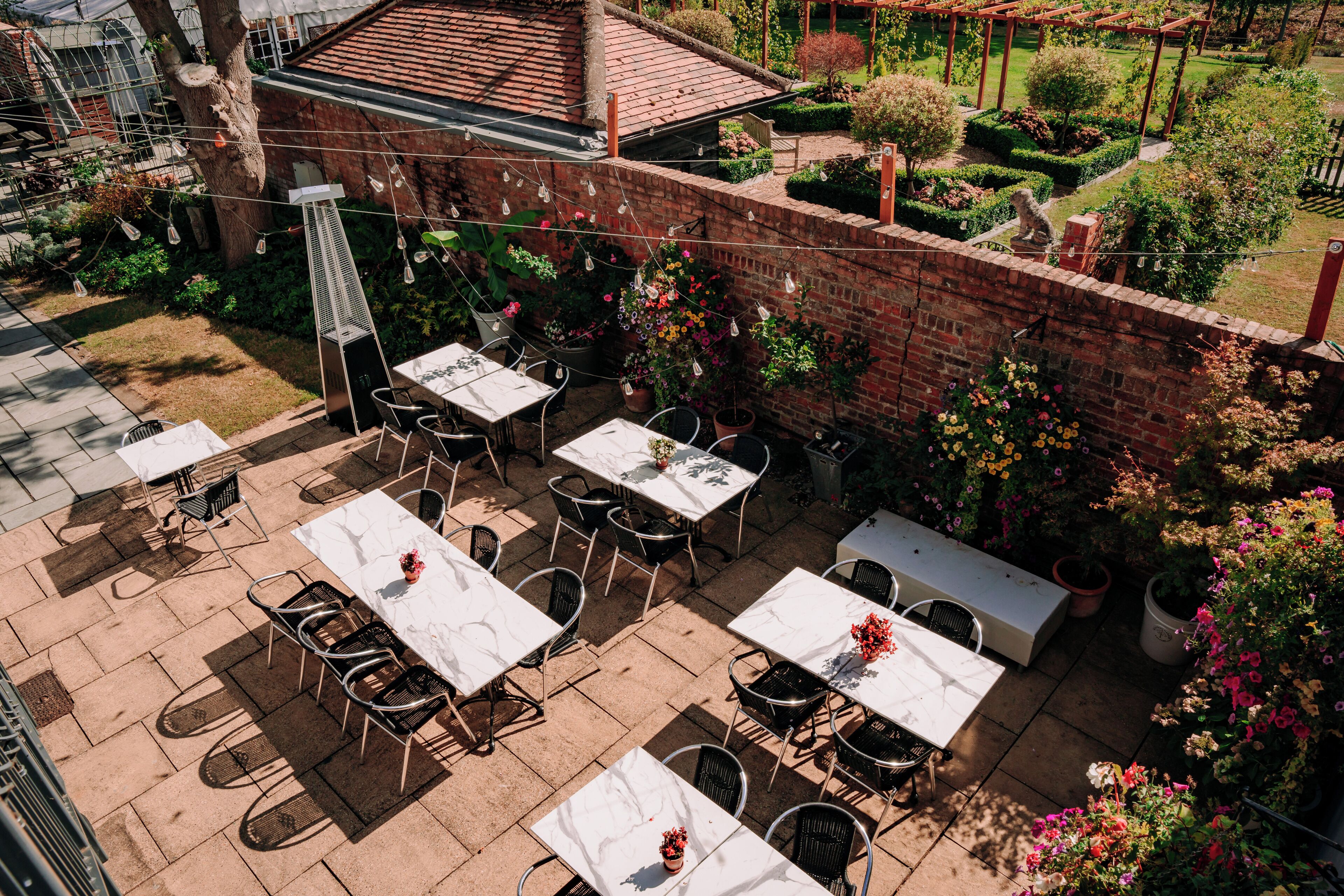 Une terrasse extérieure dotée de plusieurs tables et chaises, entourée de murs en briques et agrémentée de plantes en pot.