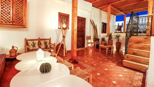 A cozy patio area with wooden furniture, a white table with a cactus, and a wooden staircase leading to an upper level. - Dar Dada Mouss (Marrakech)