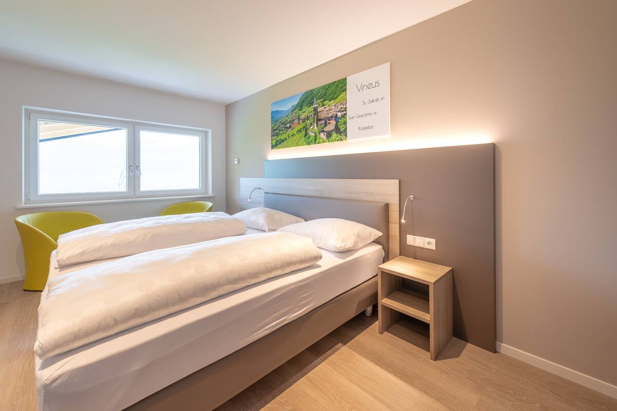 A modern bedroom with a large bed, a bedside table, a window, and a wall-mounted picture.