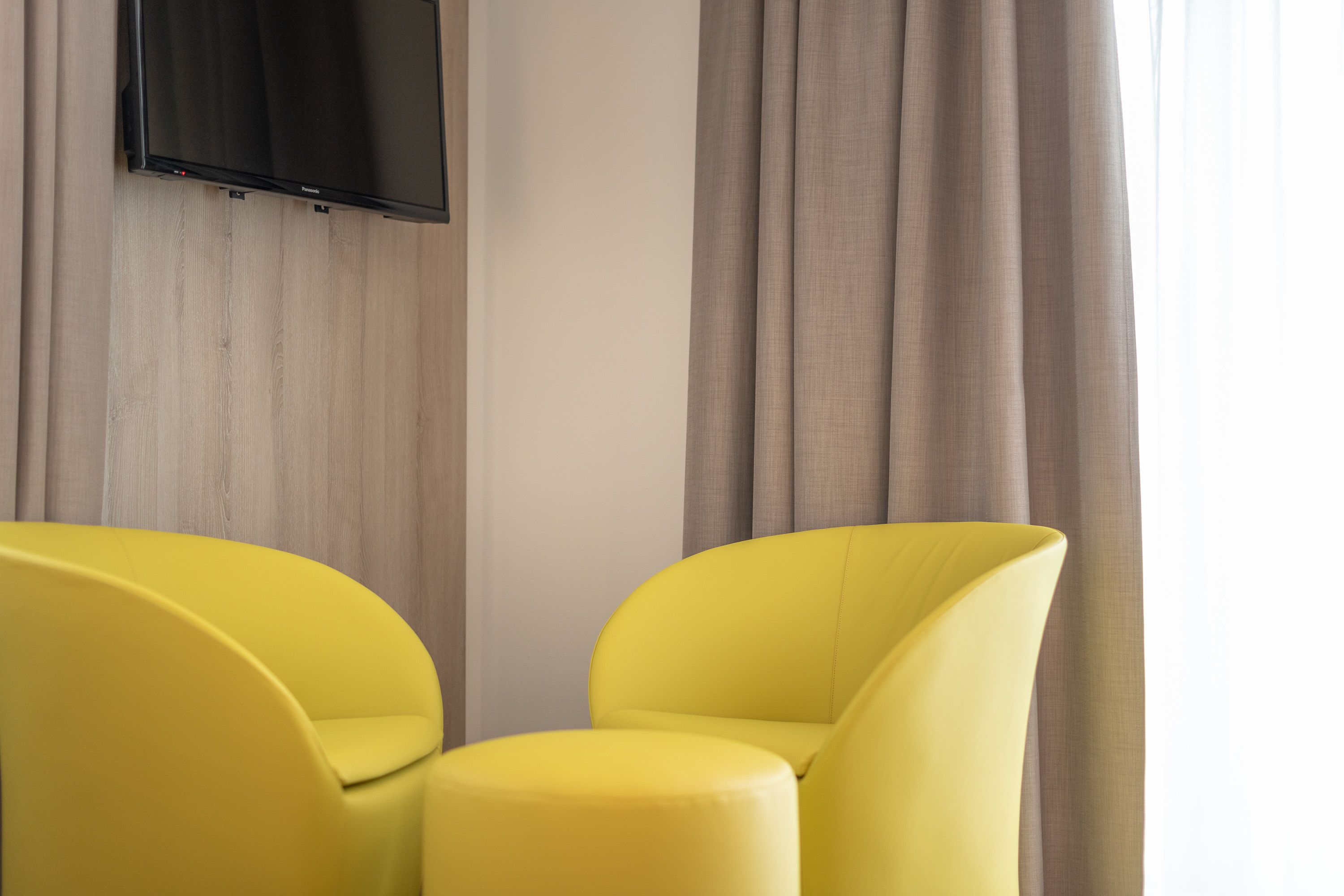 Ein Zimmer mit zwei gelben Sesseln, einem an der Wand montierten Flachbildfernseher und hellen Vorhängen.