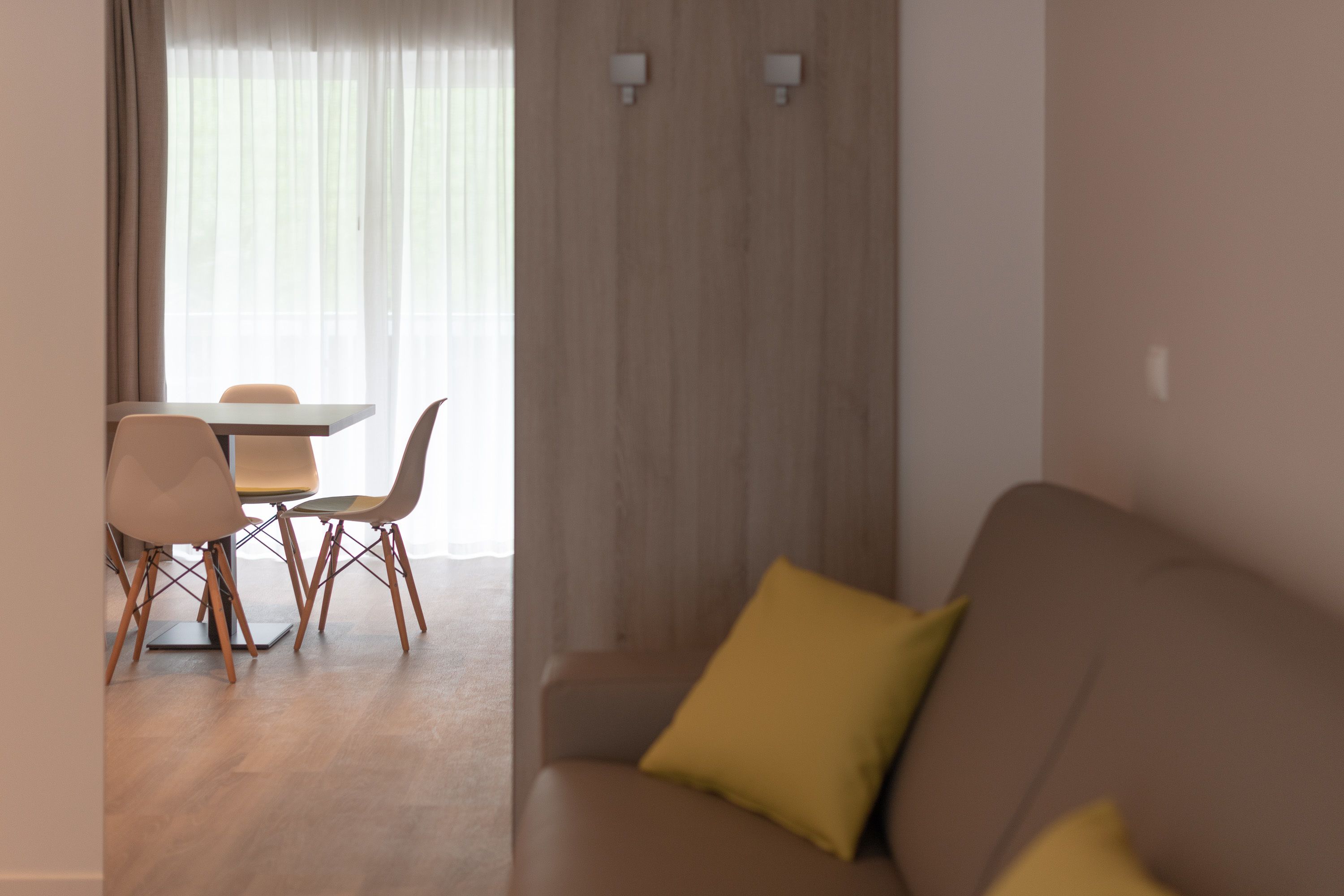Ein modernes Wohnzimmer mit einem braunen Sofa und gelben Kissen, einem Essbereich mit Tisch und Stühlen sowie einem großen Fenster mit durchscheinenden Vorhängen.