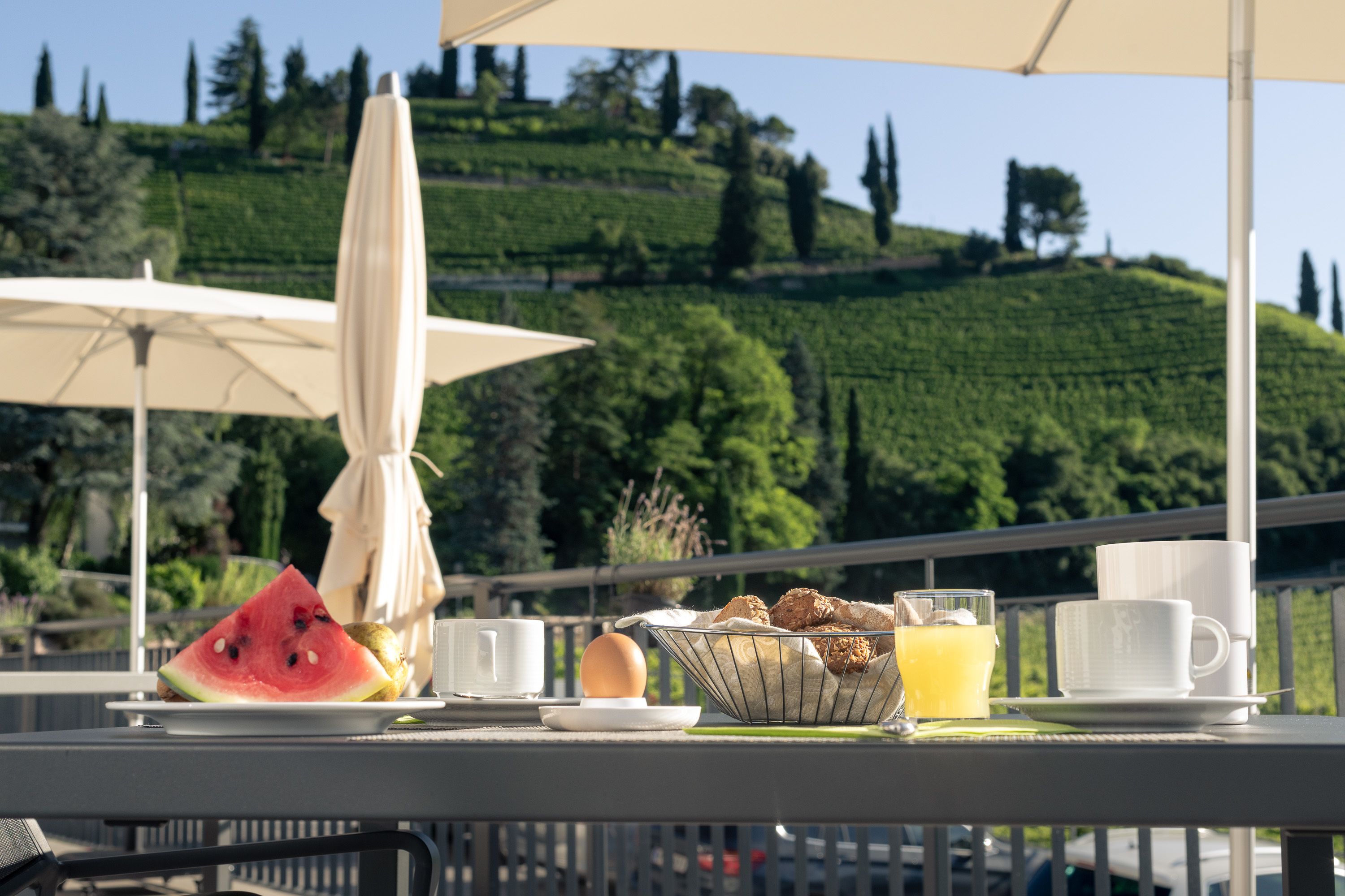 Una terrazza con un tavolo apparecchiato per la colazione, con una fetta di anguria, uova e un cesto di frutta.