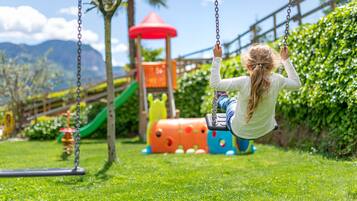 Ein Spielplatz mit Schaukeln und einer Rutsche, umgeben von viel Grün und blauem Himmel.