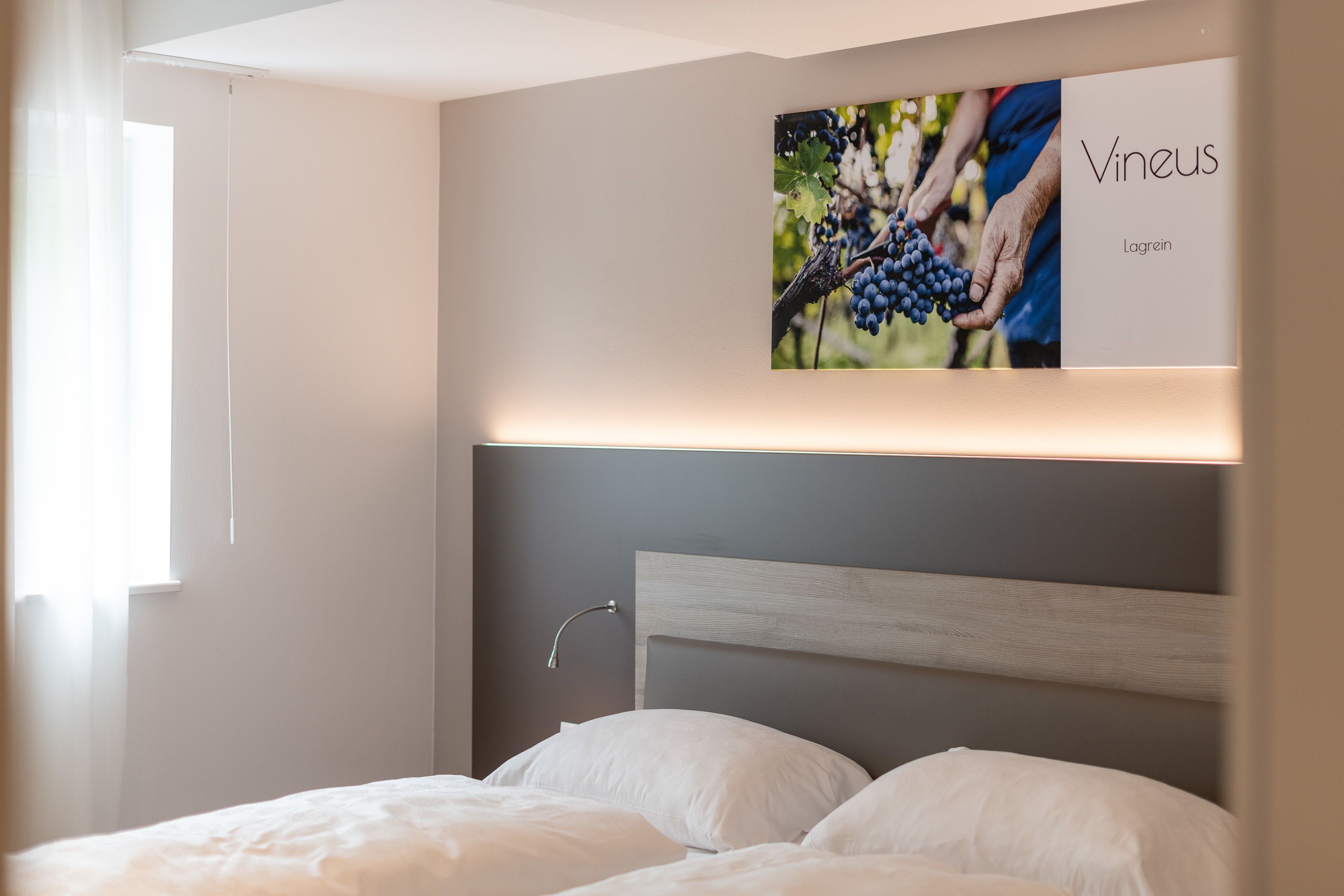 Ett hotellrum med en säng, en sänggavel och en tavla på väggen som föreställer druvor och en person.