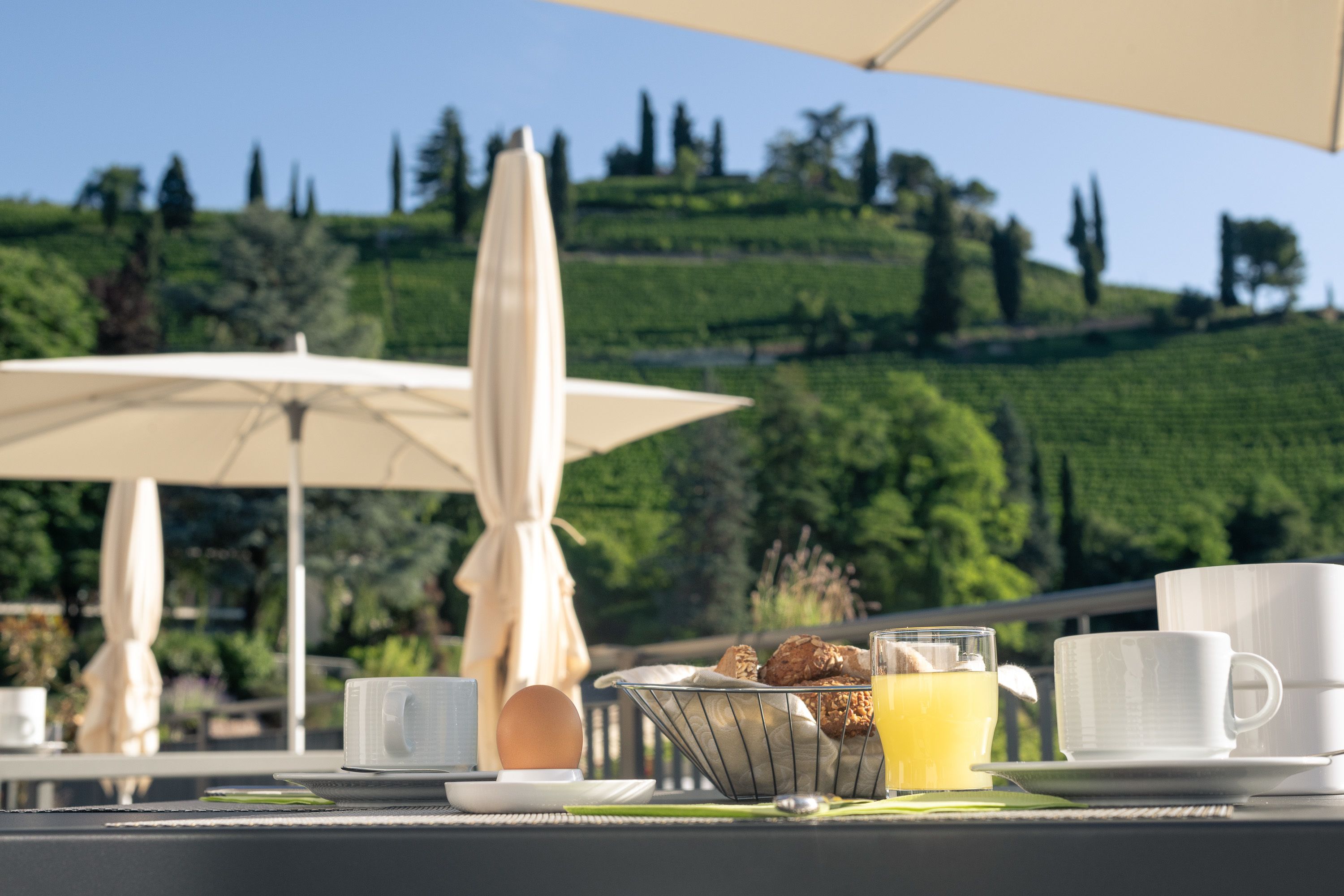 A breakfast setup with a table, a basket of pastries, a glass of orange juice, and white cups on a terrace with a scenic view of a green hill and trees.