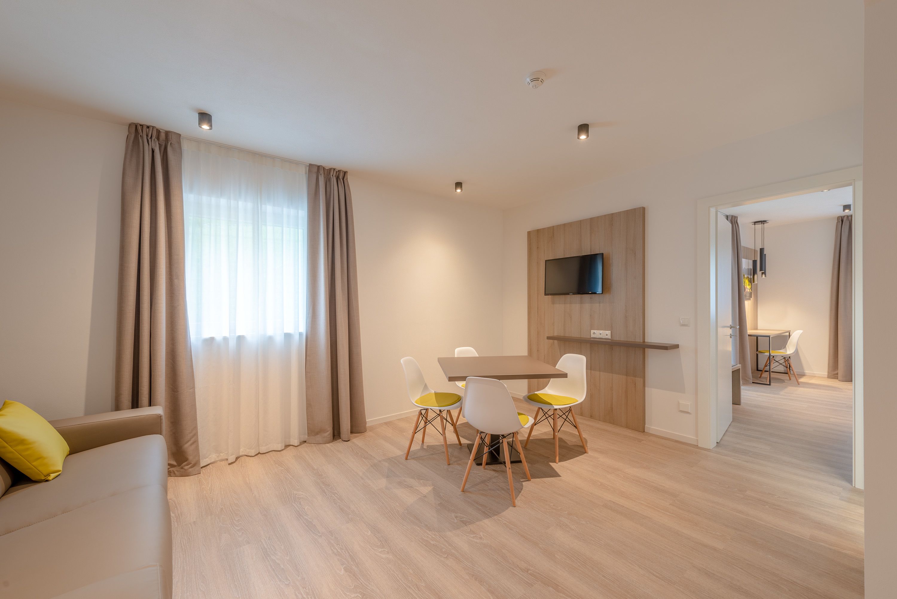 Ein modernes Wohnzimmer mit einer Couch, einem Esstisch mit Stühlen, einem an der Wand montierten Fernseher und einem Flur, der in einen anderen Raum führt.