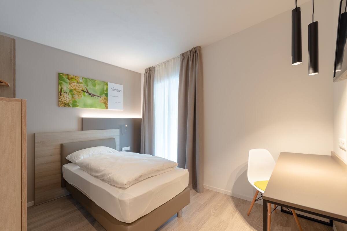 A modern hotel room with a bed, a desk, a chair, and a painting on the wall.