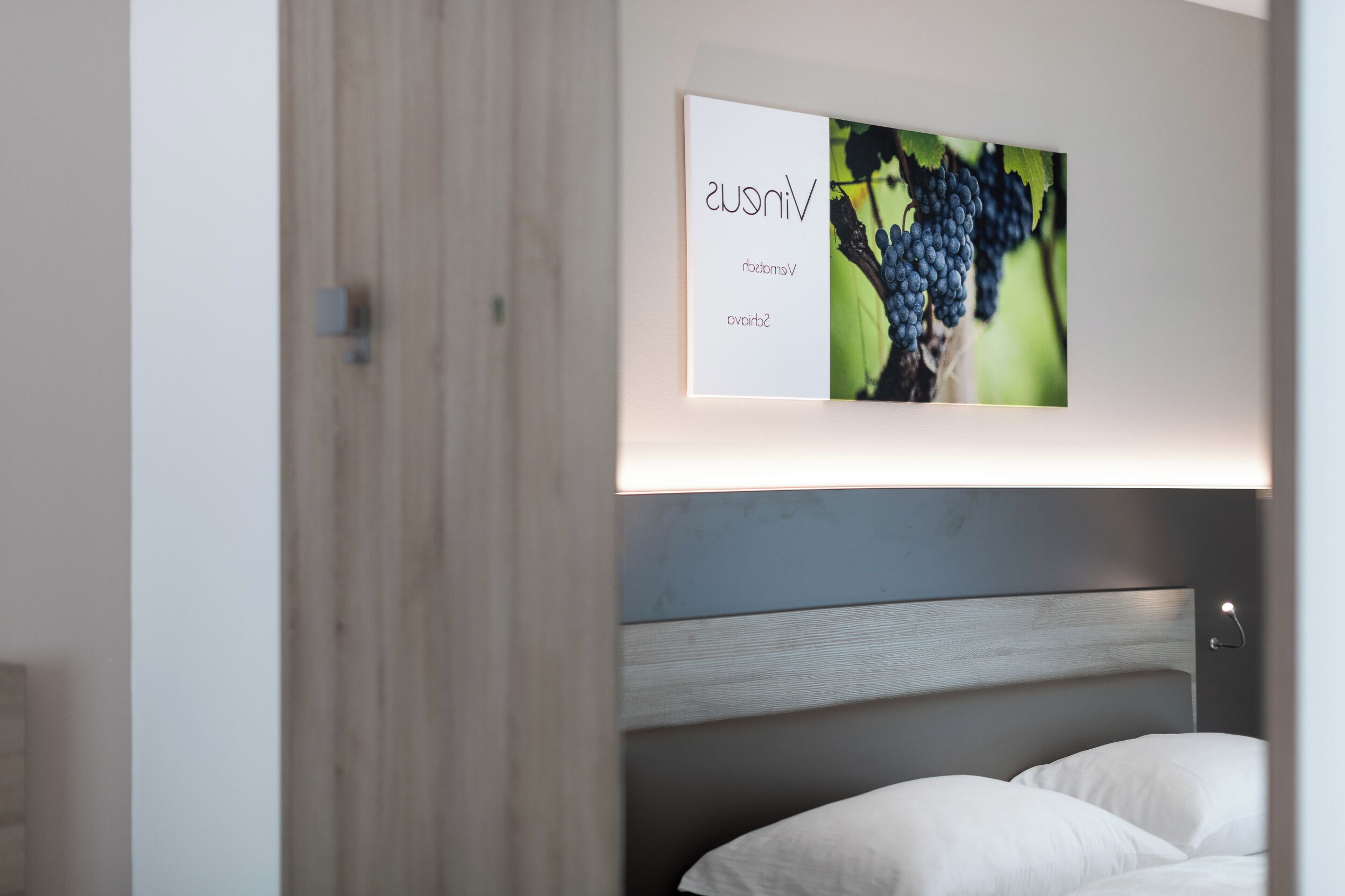 A modern bedroom with a wooden headboard, a bed with white linens, and a wall-mounted artwork depicting grapes.