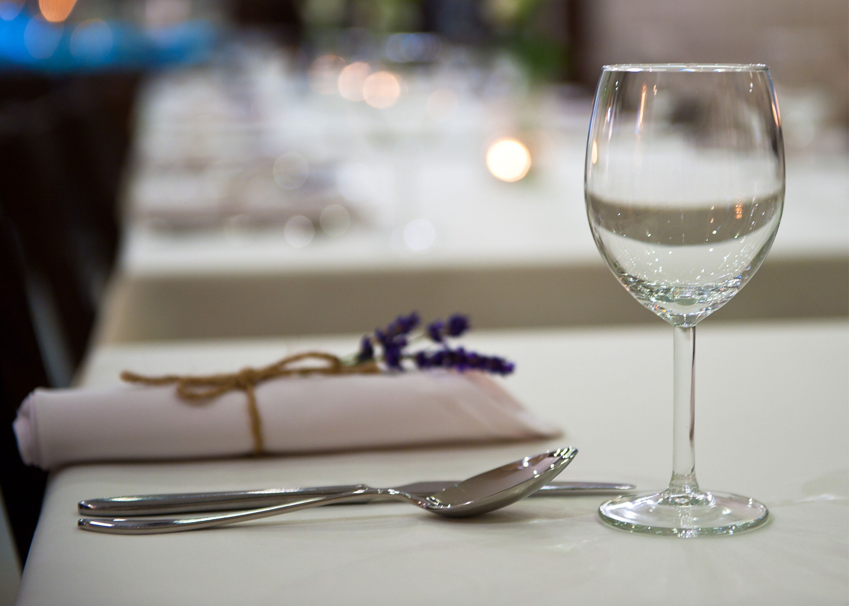 Una mesa servida con una copa de vino, una cuchara y una servilleta atada con un hilo, junto con un pequeño ramo de flores moradas.
