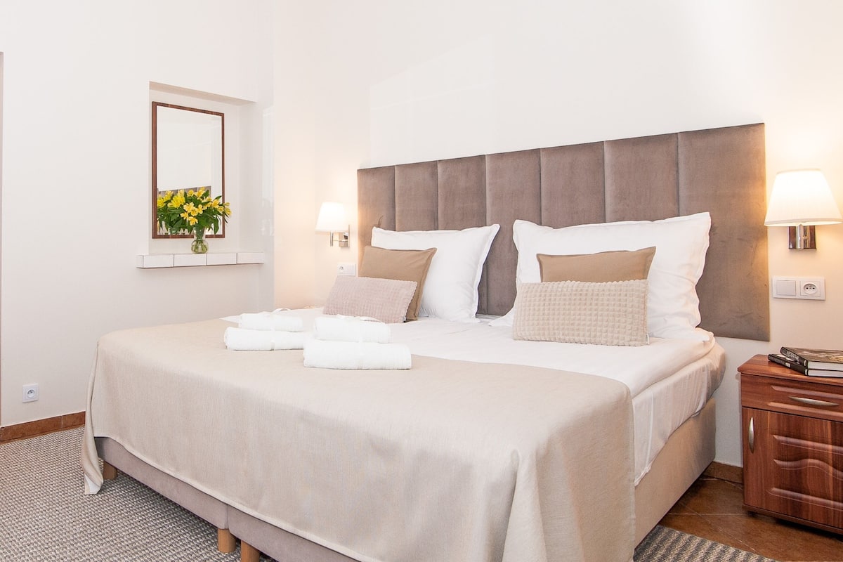 A neatly made bed with a headboard, two bedside lamps, and a bedside table with a book.