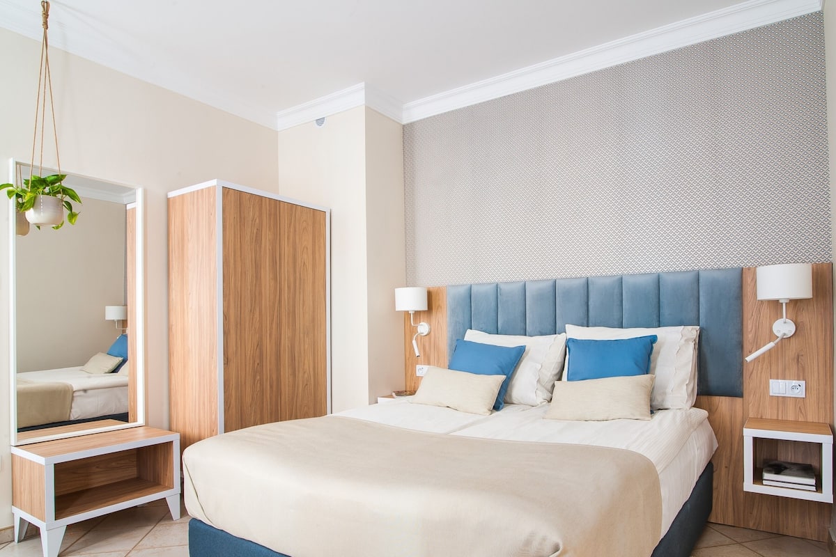 A modern bedroom with a large bed, a wardrobe, a mirror, and a nightstand with a lamp.
