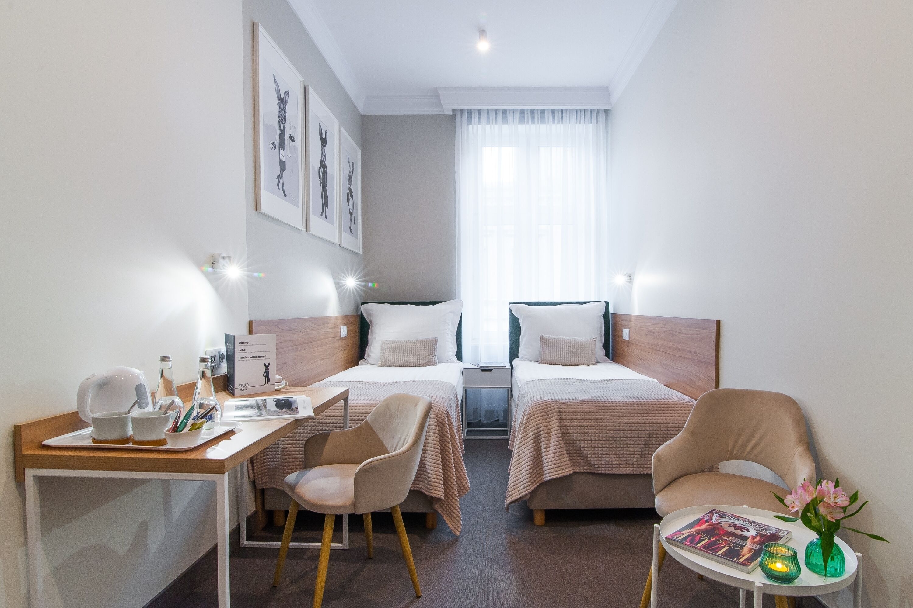 Habitación de hotel con dos camas, un escritorio con una silla, una mesita con un florero y una revista, y una ventana con cortinas translúcidas.