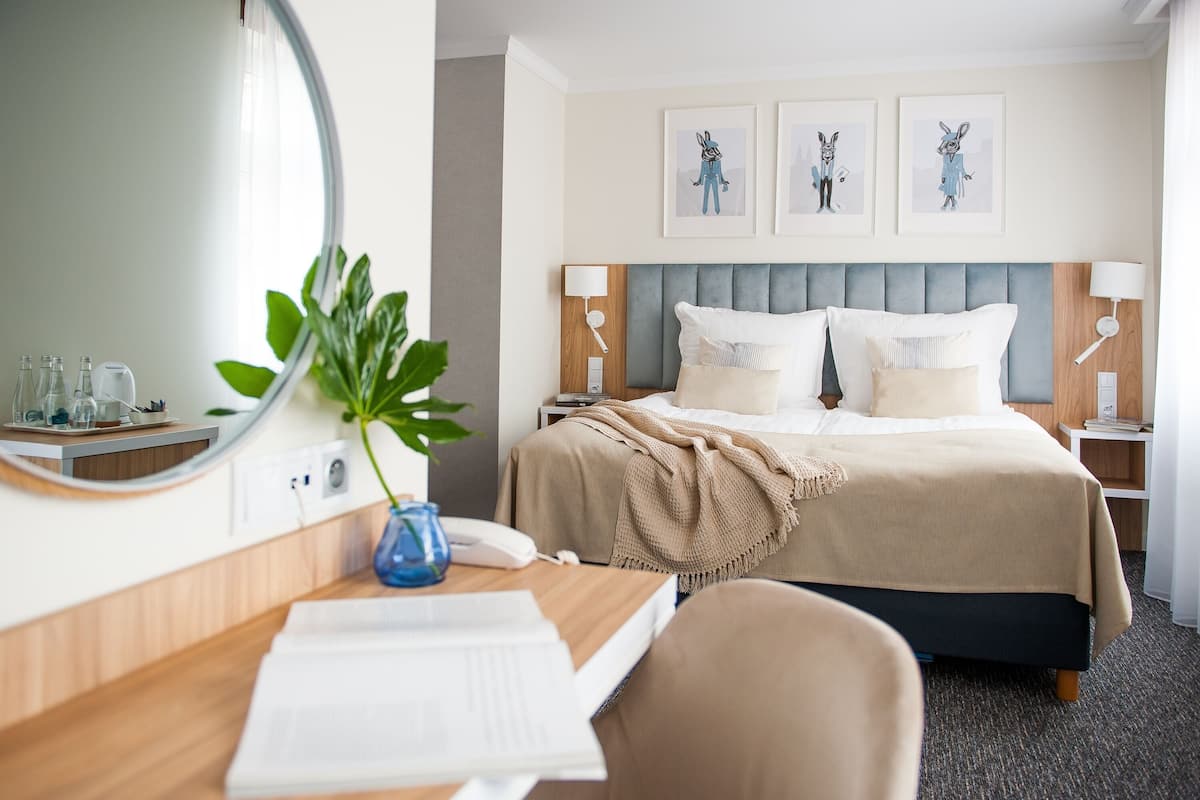 A hotel room with a bed, bedside tables, a desk with a chair, a mirror, and framed artwork on the wall.