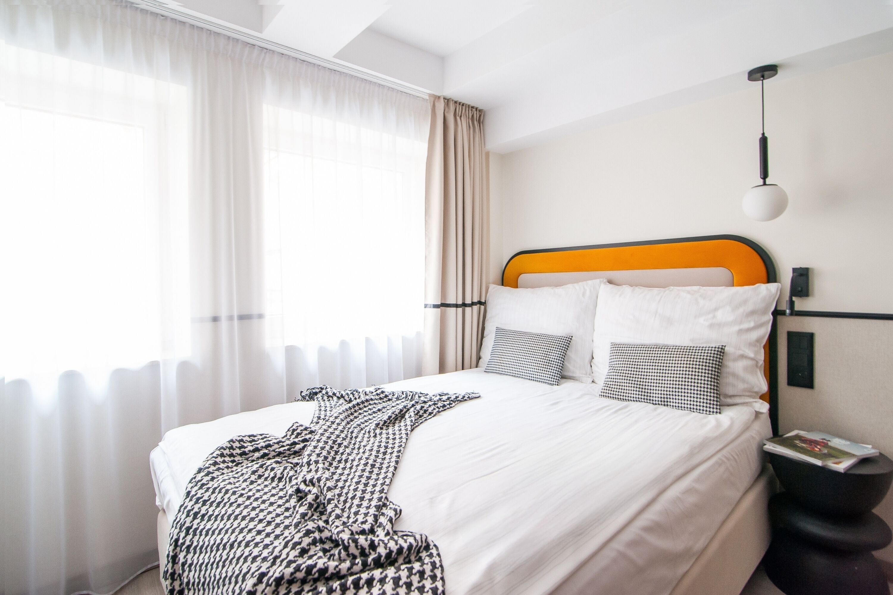 Una cama bien tendida con ropa blanca y una manta estampada, una mesita de noche negra con una revista, y una ventana con cortinas translúcidas.