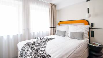 Una cama bien tendida con ropa blanca y una manta estampada, una mesita de noche negra con una revista, y una ventana con cortinas translúcidas.