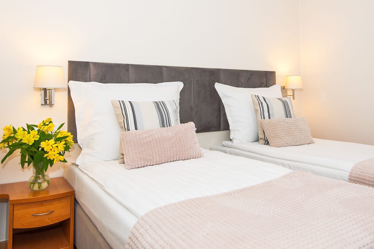 A double room with two beds, each with white linens and striped pillows, a wooden nightstand with a lamp, and a vase of yellow flowers.