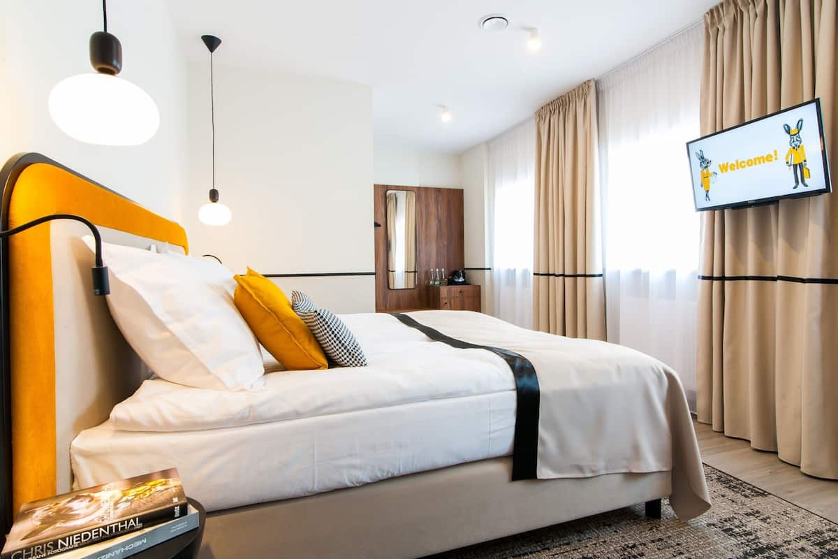 A hotel room with a bed, a TV displaying a welcome message, a nightstand, and a window with curtains.