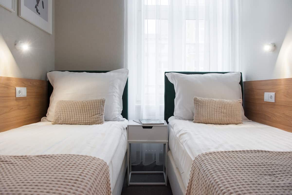 A twin room with two beds, a nightstand, a window with sheer curtains, and wall-mounted lights.