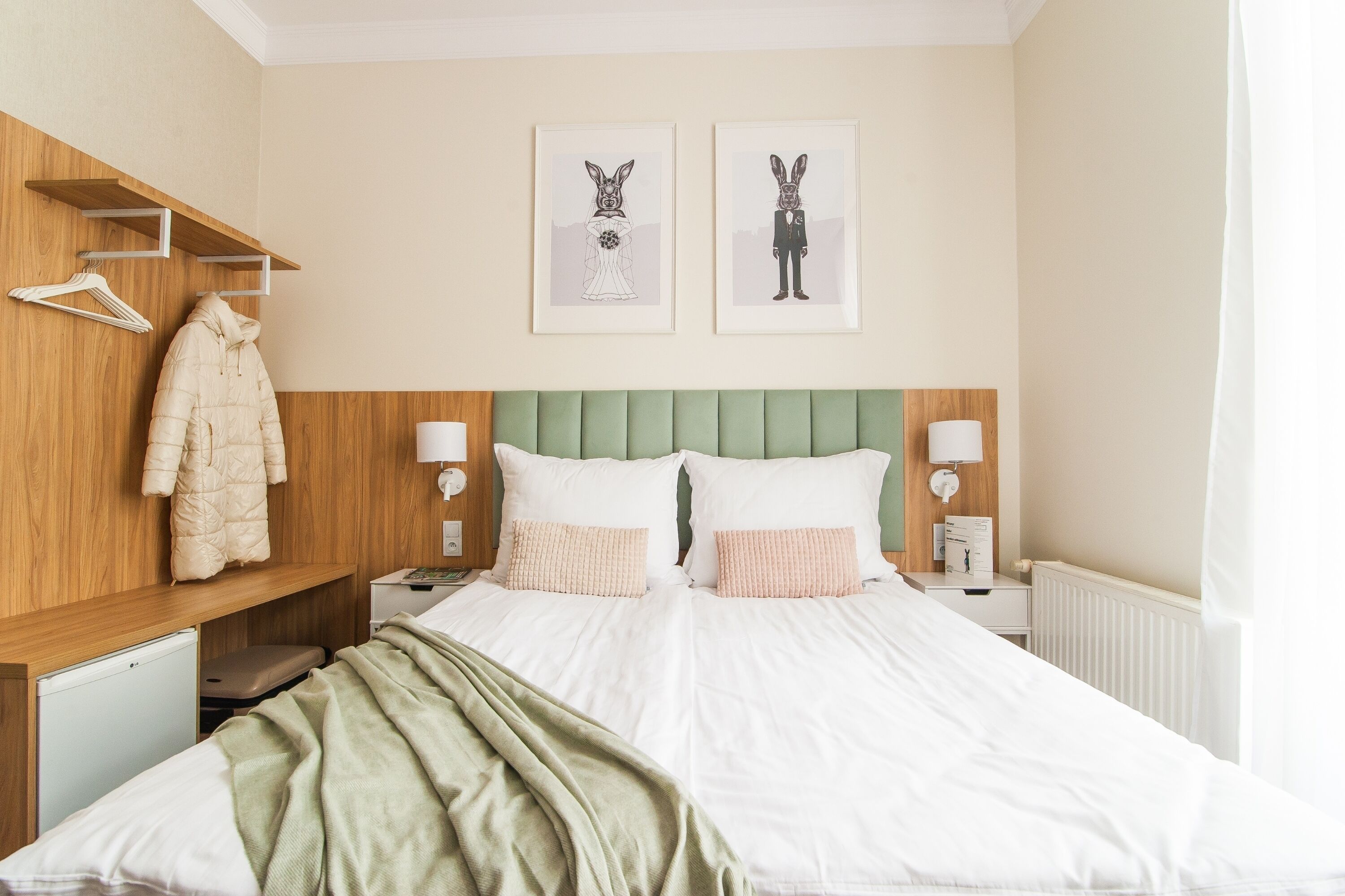 Une chambre à coucher bien rangée, avec un lit, une table de chevet, une lampe, un manteau accroché à une étagère en bois et deux tableaux encadrés au mur.