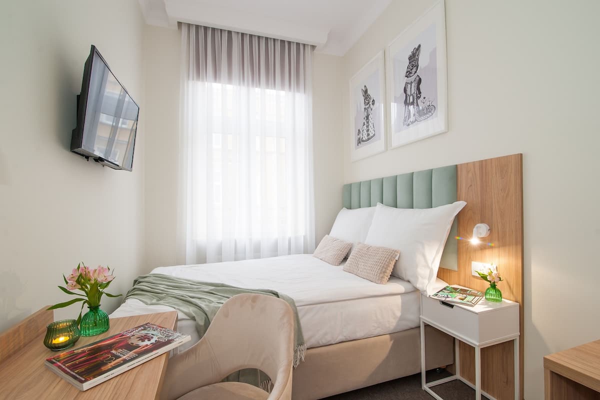 A neatly arranged hotel room with a bed, a bedside table with a lamp, a chair, a TV mounted on the wall, and artwork on the walls.