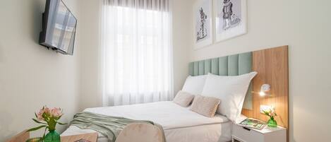 A neatly arranged hotel room with a bed, a bedside table with a lamp, a chair, a TV mounted on the wall, and artwork on the walls.