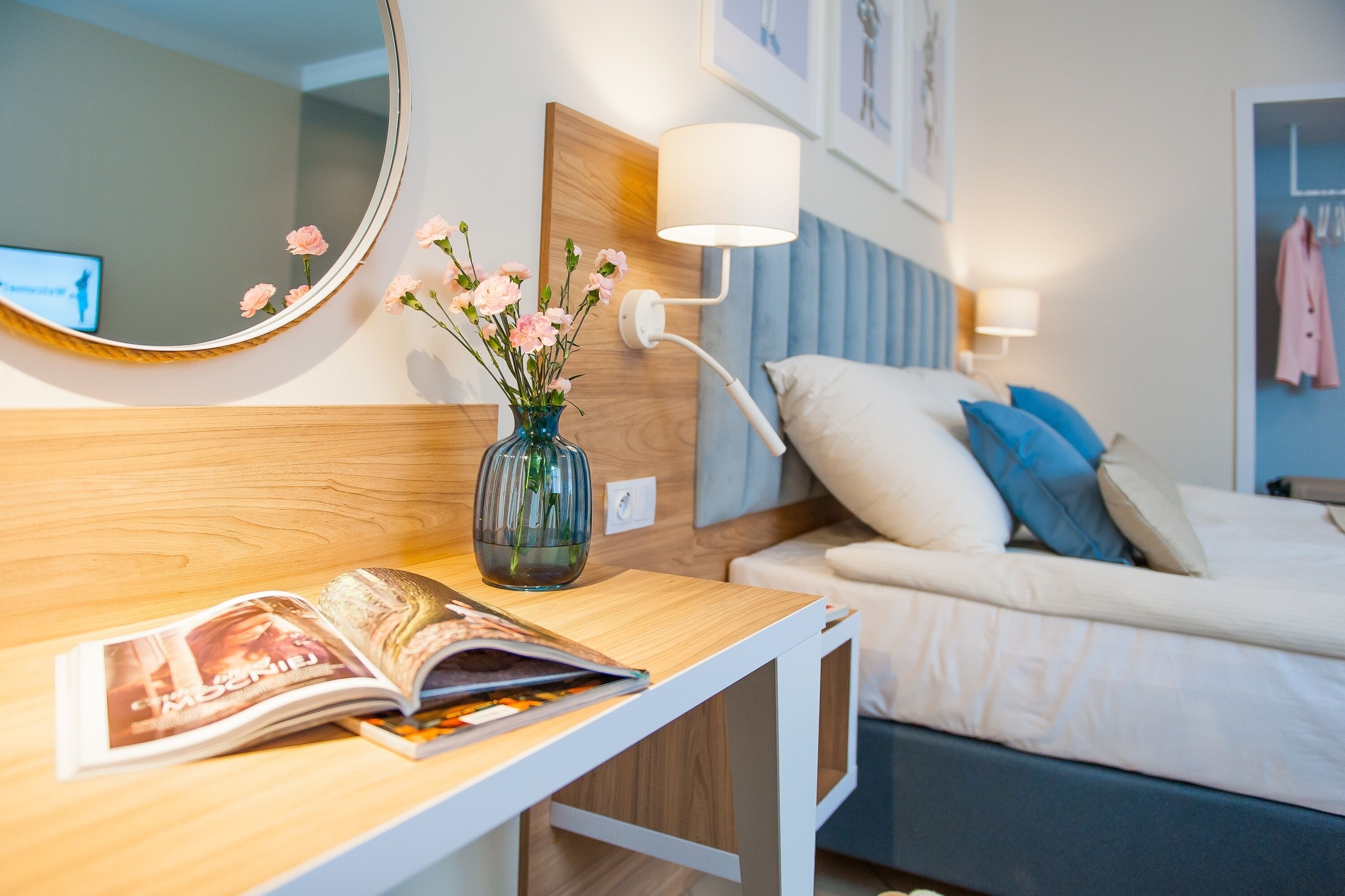 Une chambre d’hôtel moderne avec un bureau en bois, une lampe de chevet, un miroir, un vase avec des fleurs, un lit avec une literie bleue et blanche, et un tableau encadré au mur.