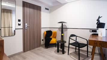 A hotel room with a wooden door, a mirror, a yellow chair, a black chair, a small round table with a vase, and a lamp.