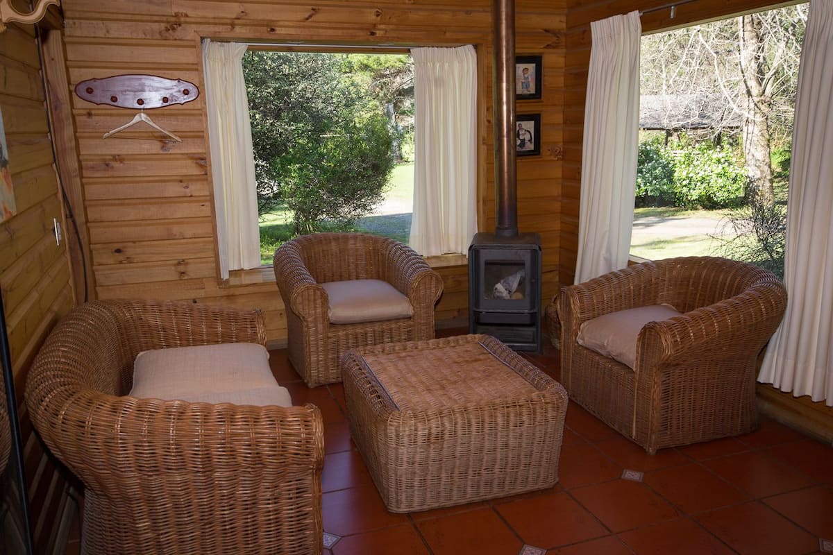 L’intérieur d’un chalet confortable, avec des meubles en osier, un poêle à bois et de grandes fenêtres ornées de rideaux.